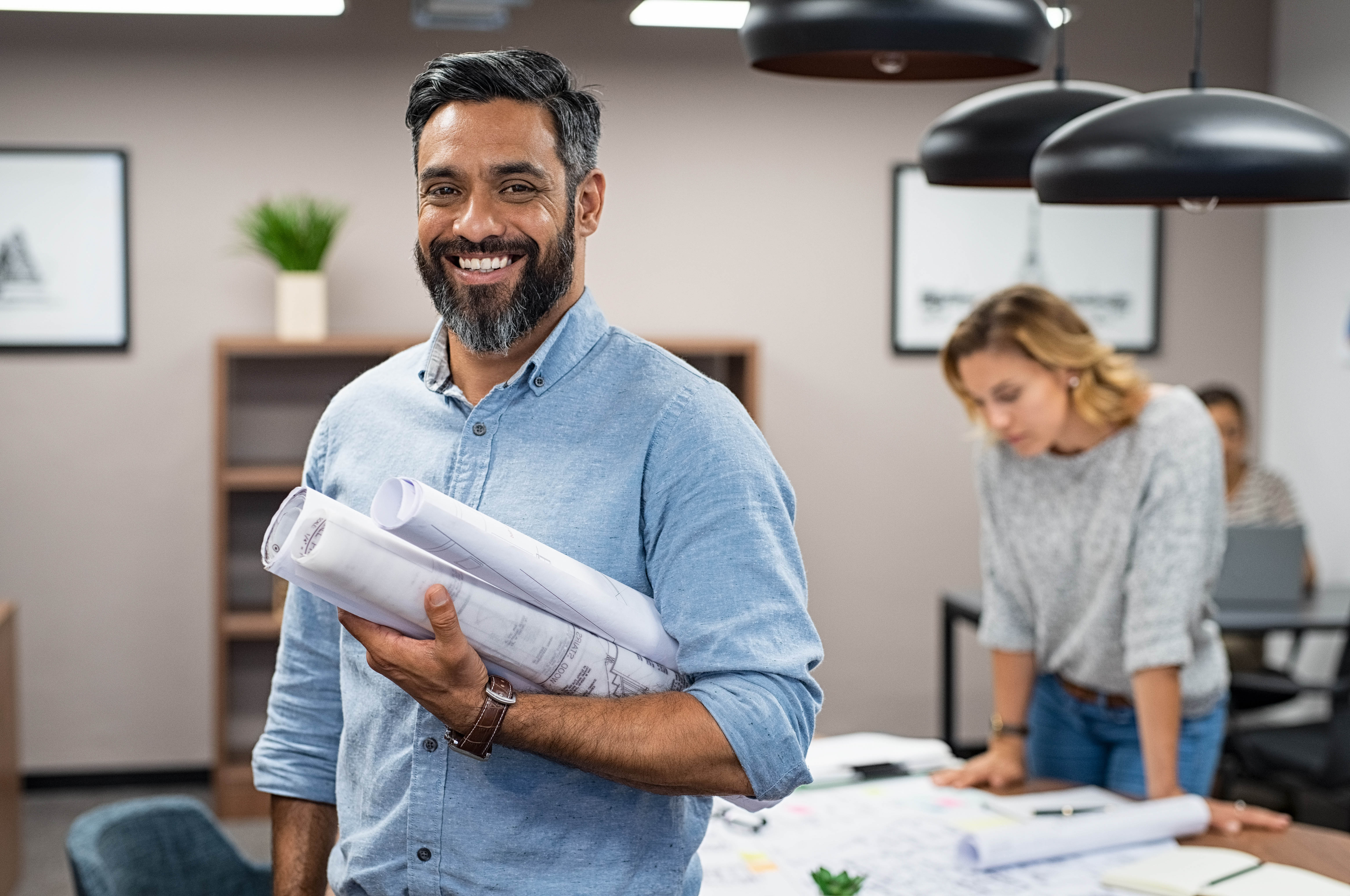 Portrait of multiethnic architect with blueprints in creative office. Mature middle eastern contractor holding roll of architectural projects while looking at camera. Happy latin man in casual.